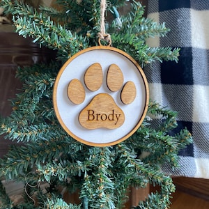 May include: A wooden Christmas ornament shaped like a paw print with the name "Brody" inscribed on it. The ornament is hanging on a small artificial Christmas tree.
