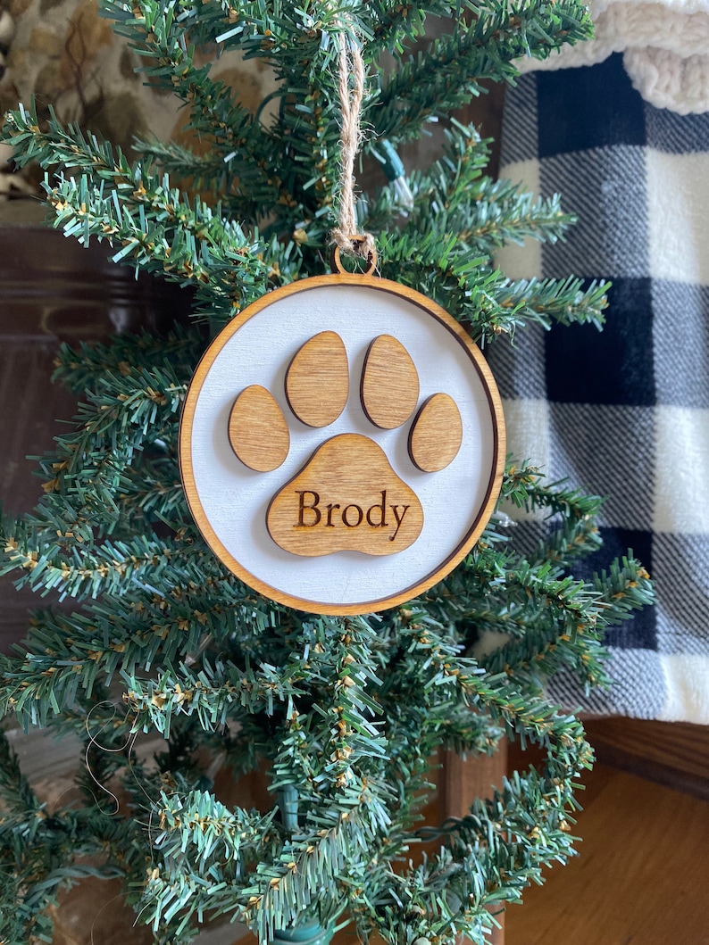 May include: A wooden Christmas ornament shaped like a paw print with the name "Brody" inscribed on it. The ornament is hanging on a small artificial Christmas tree.