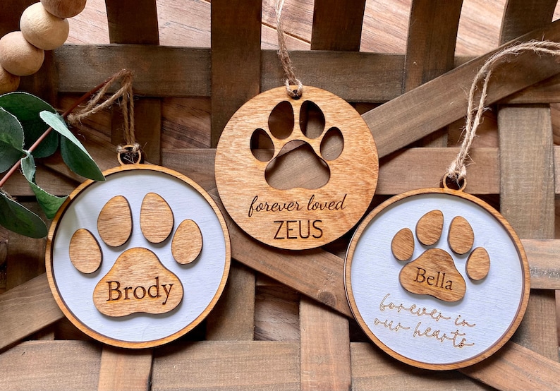 May include: Three wooden paw print ornaments with personalized names. The ornaments are brown with a white background and feature a paw print design. The text on the ornaments reads "Brody", "forever loved ZEUS", and "Bella forever in our hearts".
