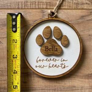 May include: A wooden ornament with a brown paw print and the name "Bella" inscribed on it. The ornament also has the text "forever in our hearts" written on it.