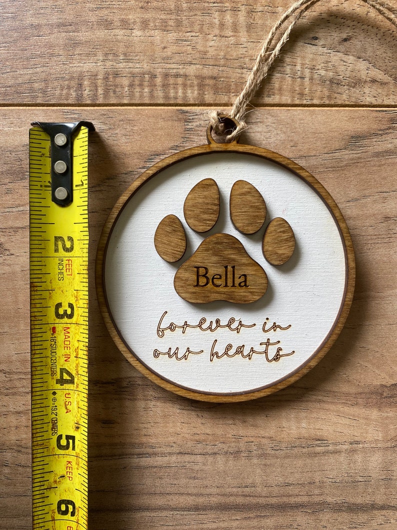 May include: A wooden ornament with a brown paw print and the name "Bella" inscribed on it. The ornament also has the text "forever in our hearts" written on it.