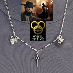 May include: Silver-toned necklace featuring a fox head, dice, and a sword pendant. The necklace is connected by a delicate chain. The Wheel of Time logo is displayed in gold and black.