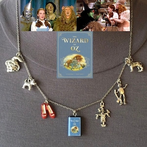 May include: Silver-toned necklace featuring charms inspired by "The Wizard of Oz." Charms include a dog, elephant, ruby slippers, a book, a scarecrow, a tin man, and a lion. The necklace is displayed on a gray background, with images from the movie in the background.