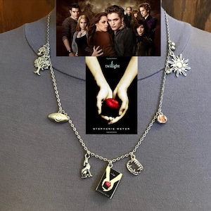May include: Silver-toned necklace featuring charms related to the Twilight saga. Charms include a tree, an eye, a wolf, a book, a set of fangs, a sun, and a pink gem. The necklace is displayed on a gray background, with the Twilight movie poster in the background.