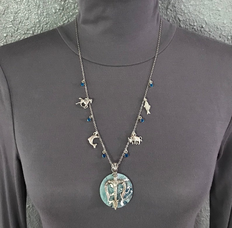 Puede incluir: Collar plateado con un colgante grande y redondo con un dise&ntilde;o azul y blanco. El collar tiene peque&ntilde;os dijes de un caballo, un delf&iacute;n, un pez y un toro, con cuentas azules. El collar se muestra sobre un cuello alto gris.