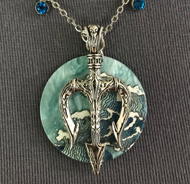 Puede incluir: Collar con colgante de tridente plateado y fondo circular con tem&aacute;tica oce&aacute;nica. El colgante presenta un dise&ntilde;o de tridente detallado. El collar incluye dos peque&ntilde;os acentos de piedras preciosas azules en la cadena.