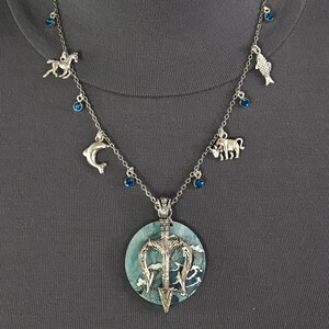 Puede incluir: Collar plateado con un colgante grande con un dise&ntilde;o de tridente sobre un patr&oacute;n de olas azules y blancas. El collar tiene dijes de caballo, delf&iacute;n, pez y toro, con detalles de piedras preciosas azules.