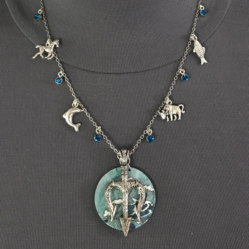 Puede incluir: Collar plateado con un colgante grande con un dise&ntilde;o de tridente sobre un patr&oacute;n de olas azules y blancas. El collar tiene dijes de caballo, delf&iacute;n, pez y toro, con detalles de piedras preciosas azules.