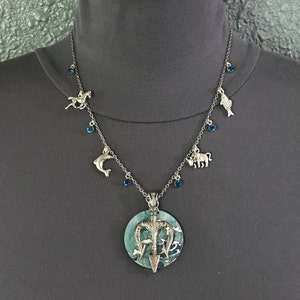 Puede incluir: Collar plateado con un colgante grande y redondo con un dise&ntilde;o de tridente y un fondo azul verdoso. El collar tiene peque&ntilde;os dijes de un caballo, delf&iacute;n, pez y toro, junto con detalles de piedras preciosas azules.