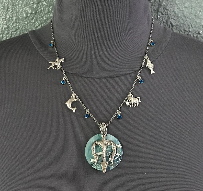 Puede incluir: Collar plateado con un colgante grande y redondo con un dise&ntilde;o de tridente y un fondo azul verdoso. El collar tiene peque&ntilde;os dijes de un caballo, delf&iacute;n, pez y toro, junto con detalles de piedras preciosas azules.