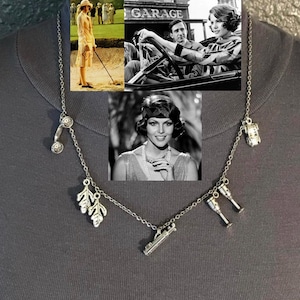 May include: Silver-toned necklace with multiple charms, including a telephone, leaves, a car, a gun, and champagne flutes. The necklace is displayed against a dark background, with vintage black and white images in the background, evoking a classic, retro theme.