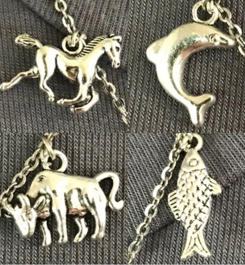 Puede incluir: Cuatro collares con colgantes plateados. Los colgantes incluyen un caballo corriendo, un delf&iacute;n, un toro y un pez. Cada colgante est&aacute; unido a una delicada cadena plateada. Los collares se muestran sobre un fondo de tela gris.