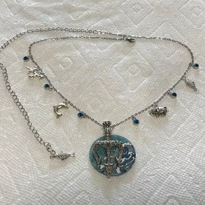 Puede incluir: Collar plateado con un colgante redondo con un dise&ntilde;o de olas azules y blancas y un tridente. El collar tiene peque&ntilde;os dijes de criaturas marinas y gemas azules. La cadena es ajustable.