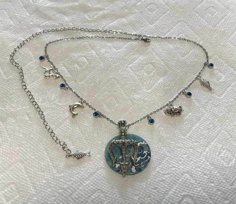 Puede incluir: Collar plateado con un colgante redondo con un dise&ntilde;o de olas azules y blancas y un tridente. El collar tiene peque&ntilde;os dijes de criaturas marinas y gemas azules. La cadena es ajustable.