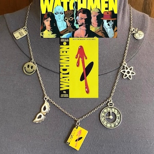 Può includere: Collana color argento con vari ciondoli, tra cui una faccina sorridente, una maschera, un orologio e un ciondolo a forma di libro con la scritta "WATCHMEN". Sullo sfondo, una copertina di fumetto con il titolo "WATCHMEN".