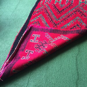 May include: A folded red and green rug with a geometric pattern. The rug has black fringe and is folded in half.