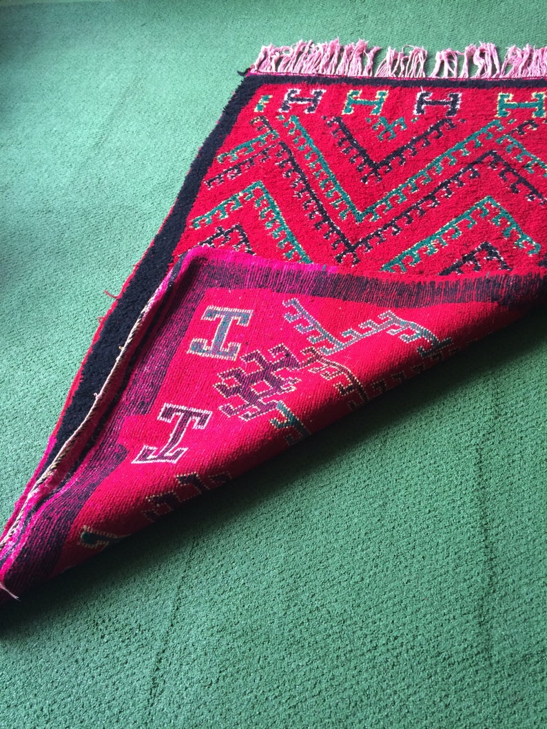 May include: A folded red and green rug with a geometric pattern. The rug has black fringe and is folded in half.