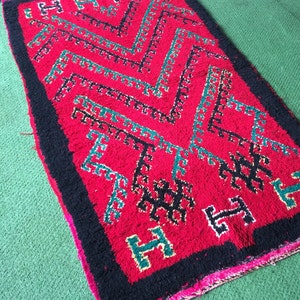May include: A red rug with a black and green geometric pattern. The rug has a fringe border.