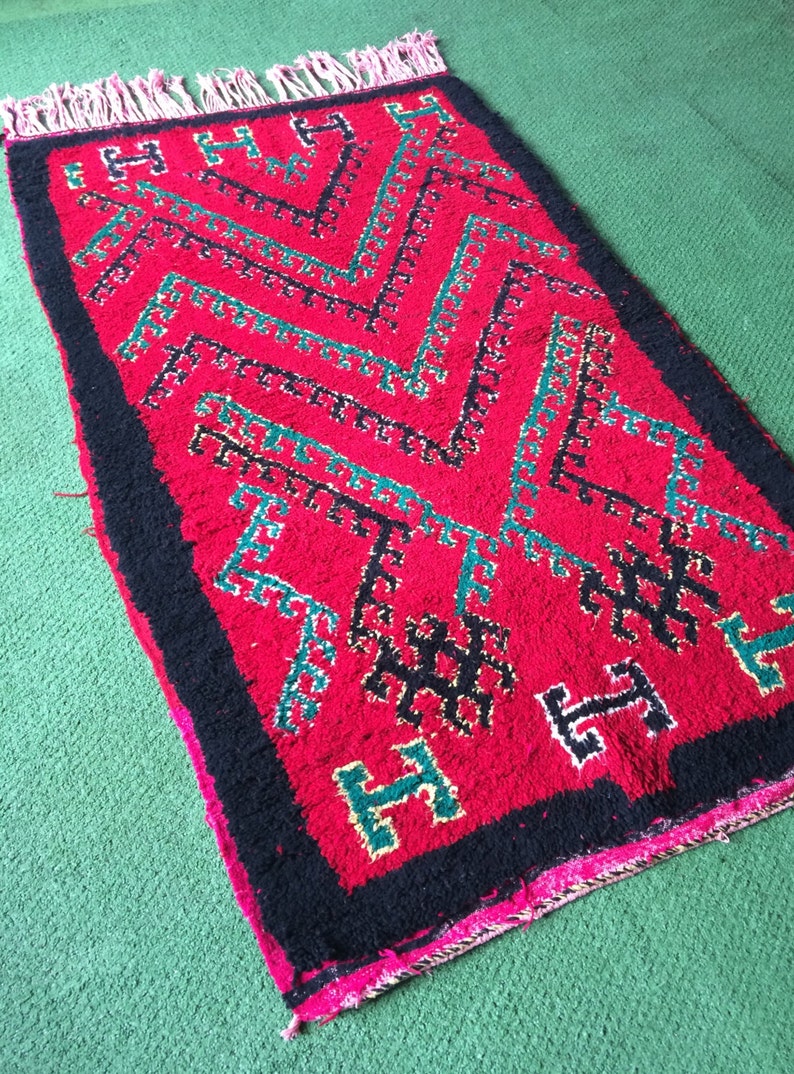 May include: A red rug with a black and green geometric pattern. The rug has a fringe border.