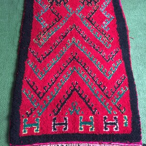 May include: A red, black, and green Moroccan rug with a geometric pattern and fringe. The rug has a black border and a pink fringe.