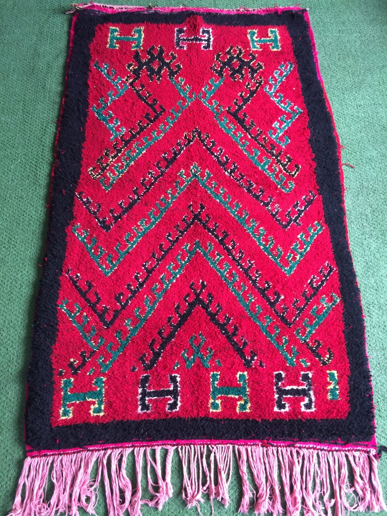 May include: A red, black, and green Moroccan rug with a geometric pattern and fringe. The rug has a black border and a pink fringe.