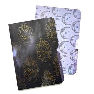 May include: Two paper file folders. The front folder is black with a repeating gold-colored palmistry hand pattern featuring sun and moon symbols. The back folder is light purple with a repeating pattern of dark gray crescent moons and stars.