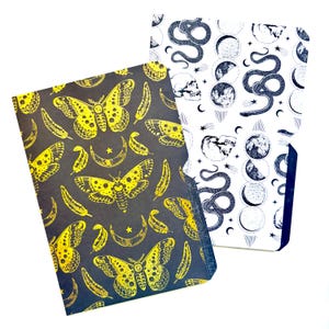 May include: Two decorative folders. One folder has a black background with yellow moth, feather, crescent moon, and star patterns. The other folder has a white background with a black snake, moon phase, skull, and star patterns. The folders are for stationery.