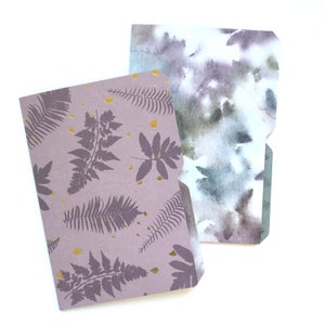 May include: Two file folders with different designs. One folder has a watercolor pattern in shades of gray and blue. The other folder has a purple background with a gold foil pattern of ferns.