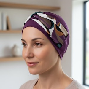 Puede incluir: Un gorro tipo turbante de color púrpura oscuro con un adorno estampado. El adorno presenta un diseño de formas blancas, marrones, verdes y negras. El gorro está hecho de un material suave y está diseñado para ser cómodo.