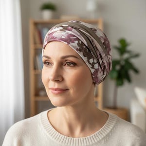 Könnte beinhalten: Eine Frau trägt eine Kopfbedeckung mit einem Blumenmuster in Lila-, Weiß- und Brauntönen. Die Kopfbedeckung ist im Turbanstil mit einem einfarbigen braunen Band gewickelt. Die Frau trägt einen weißen Pullover.