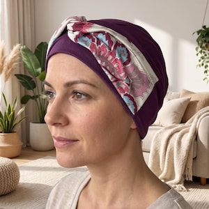 May include: A deep purple turban-style head covering featuring a wide, twisted front band. The band is decorated with a multi-colored floral pattern in shades of pink, light blue, and white, contrasting against the solid dark purple fabric base.