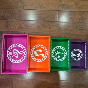 Puede incluir: Un juego de cuatro bandejas rectangulares de madera encajables en colores rosa, naranja, verde y morado. Cada bandeja presenta un diseño circular blanco recortado con un borde en forma de corazón y un símbolo de nota musical blanco.