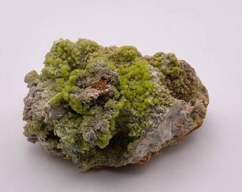 May include: A cluster of green and grey minerals on a rough, brown rock. The green minerals are clustered together in a dense, rounded shape.