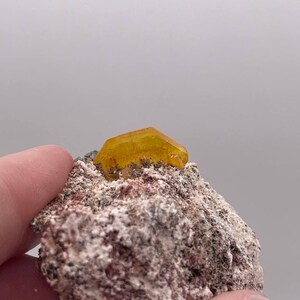 May include: A small, yellow, translucent crystal sits atop a rough, gray and brown rock. The crystal is a hexagonal prism with sharp edges and a pointed tip.