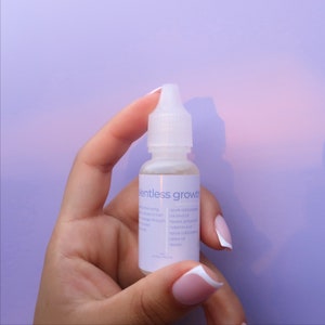 May include: A clear glass bottle with a dropper top, labeled "entless growth". The bottle contains a light yellow liquid and is held in a hand with pink and white nail polish.