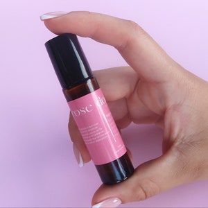 Op de afbeelding: Een roze en bruine rollerfles met de tekst "rose dew" op het etiket. De fles wordt vastgehouden door een hand met witte nagellak.