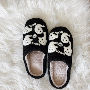 May include: A pair of black fuzzy slippers with white ghost and "BOO" designs. The slippers have a soft, beige interior and are set against a white, fluffy background. These cozy slippers are perfect for lounging at home.