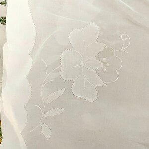 Pode incluir: Tecido branco transparente com um delicado desenho floral bordado. O bordado apresenta uma flor grande com uma videira e folhas.