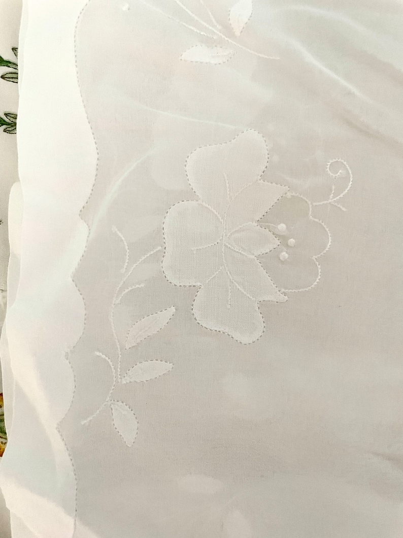 Pode incluir: Tecido branco transparente com um delicado desenho floral bordado. O bordado apresenta uma flor grande com uma videira e folhas.
