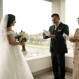 May include: A bride in a white wedding dress with a long veil stands with her father and mother. The father is holding a white envelope and the mother is holding a white envelope. The bride is holding a bouquet of white flowers.