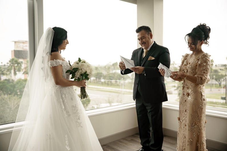 May include: A bride in a white wedding dress with a long veil stands with her father and mother. The father is holding a white envelope and the mother is holding a white envelope. The bride is holding a bouquet of white flowers.