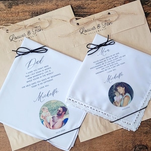 May include: Two white handkerchiefs with black text and photos, each tied with a black ribbon. One says "Dad" and the other "Mom." Both have a photo and the date "03.21.2026". They rest on brown paper bags with the text "Loaves & Fish Fellowship".