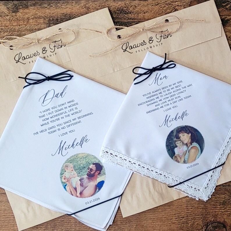 May include: Two white handkerchiefs with black text and photos, each tied with a black ribbon. One says "Dad" and the other "Mom." Both have a photo and the date "03.21.2026". They rest on brown paper bags with the text "Loaves & Fish Fellowship".
