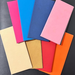 Puede incluir: Una colección de cuadernos coloridos en varios tonos, incluyendo rosa, azul, rojo y dorado. Los cuadernos están dispuestos en forma de abanico, mostrando sus vibrantes cubiertas. La imagen está tomada desde una perspectiva cenital.