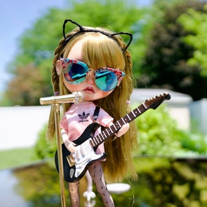 May include: A doll with blonde hair wearing black cat ears, sunglasses, a pink shirt with the Adidas logo, black fishnet tights, and brown boots, holding a black electric guitar and a microphone.