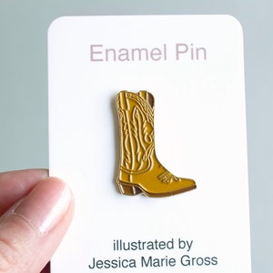 Puede incluir: Un pin de esmalte dorado con forma de bota de vaquero. El pin está sobre una tarjeta blanca que dice "Enamel Pin" en la parte superior. La tarjeta también incluye el texto "illustrated by Jessica Marie Gross" e información de redes sociales.