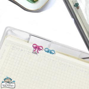 Peut inclure: Un classeur en plastique transparent avec un intercalaire en papier quadrillé. Deux petits autocollants en forme de nœud sont placés en haut de l'intercalaire, l'un rose et l'autre bleu. Le texte "My Little Blue Monkey" est en bas de l'intercalaire.