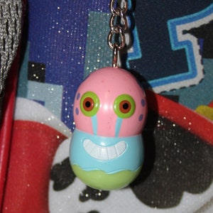 May include: A pink, blue, and green cartoon character keychain hanging from a silver chain. The keychain is attached to a red and blue backpack with a Paw Patrol design. The keychain has a smiling face.