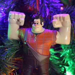 May include: A plastic ornament of a cartoon character with brown hair, a red and white plaid shirt, and brown overalls. The character is flexing both arms and has a big smile on his face. The ornament is hanging on a green Christmas tree.