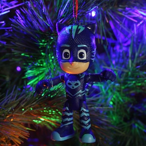 May include: A blue and black plastic ornament of a superhero character with a cat mask. The ornament is hanging on a green Christmas tree with colorful lights.